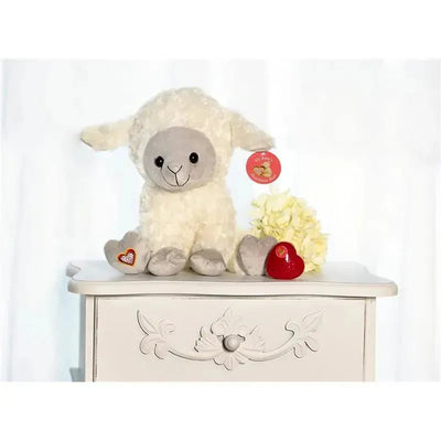 Plush lamb toy with gray face and white fur, sitting on a table next to a red heart and white flowers.