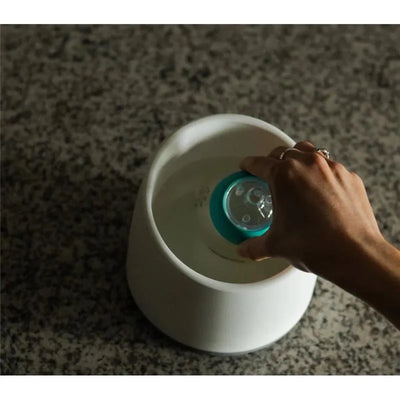 Nanobebe teal breastmilk bottle warming bowl with a hand placing a bottle into warm water.