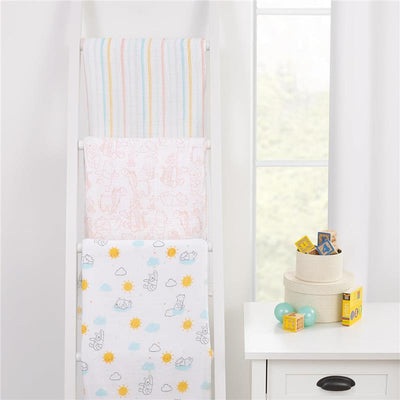 Three Nojo muslin swaddles featuring colorful designs: striped, Winnie the Pooh characters, and sun/cloud patterns displayed on a white rack.