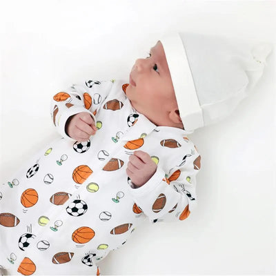 Baby wearing a Noomie zip-up footie decorated with various sports balls, paired with a white hat.