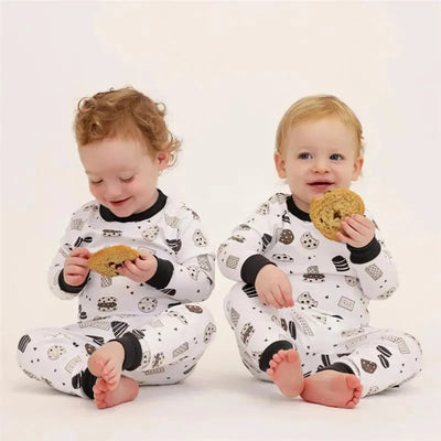 Two toddlers in cookie-patterned long sleeve pajamas sit on a light background, each holding a cookie.