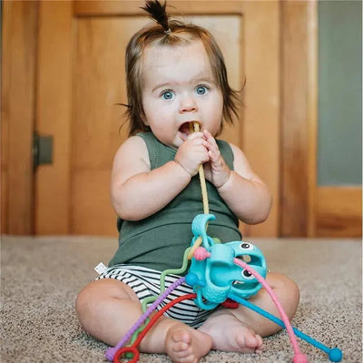 Baby sitting on carpet, chewing on a pull string of a Nuby silicone octopus toy, which features colorful tentacles.