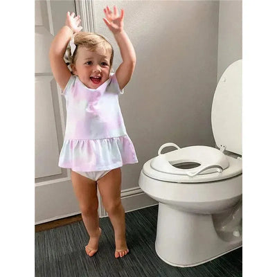 Child in a colorful top celebrates while standing next to a white toilet with a potty seat attachment.