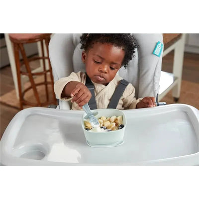 Child using a Nuk For Nature Pretensil set to eat fruit in a high chair, featuring a light green bowl and gray spoon.