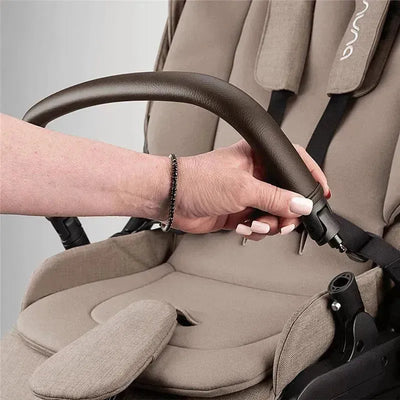 A hand adjusts the brown handle of the Nuna SWIV™ Stroller, featuring a beige fabric seat and safety harness.