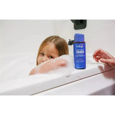 Child in a bubble bath looks towards the camera with a bottle of Oilogic Kids Cold & Cough Vapor Bath nearby.