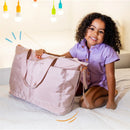 Smiling girl in a purple outfit holding a Primo Passi pink diaper bag on a bed, with colorful lights in the background.