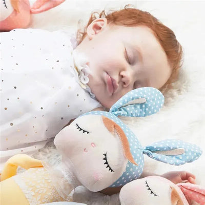 Sleeping baby girl with red hair in white dress cuddling a blue polka dot plush doll and surrounded by other dolls.