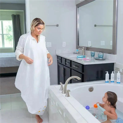 Hooded white baby towel with apron shown in a bathroom, while a woman approaches a baby in the bathtub.