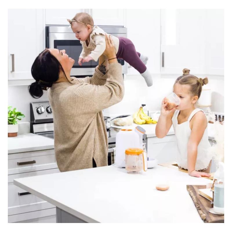 A mother holds a baby above her while a young girl drinks from a cup near a Quark Quook baby food processor on the kitchen counter.