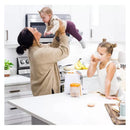 A mother holds a baby above her while a young girl drinks from a cup near a Quark Quook baby food processor on the kitchen counter.