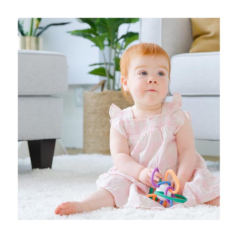 Baby girl in a pink dress sits on a soft rug, holding a colorful Quark Thiingy teething toy with various rings.