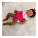 Reborn baby doll Emily, African American, with closed eyes, wearing a pink outfit and a bow, resting on a fluffy white surface.