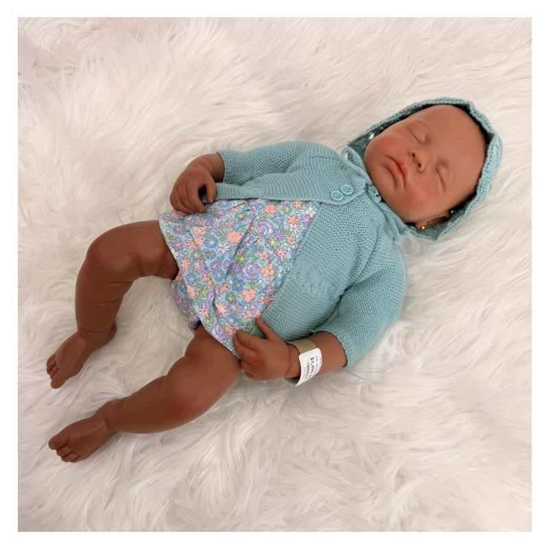 Reborn baby doll, African American, wearing a blue cardigan and floral dress, resting on a fluffy white surface.