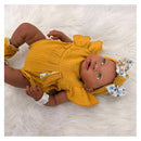 Reborn baby doll with African American features, wearing a yellow outfit and matching bow, posed on soft white fabric.