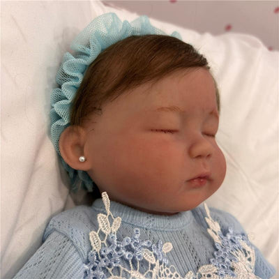 Reborn baby doll with a full silicone body, featuring a peaceful expression, blue dress, and a blue ruffled headband.
