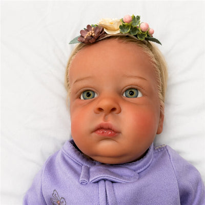 Reborn baby doll named Ana Laura with white vinyl, blonde hair, green eyes, and a floral headband, wearing a lavender outfit.