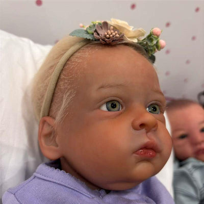 Reborn baby doll with a detailed, lifelike face, light blonde hair, a floral headband, and wearing a purple outfit.