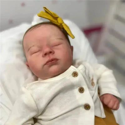 Reborn baby doll with a white vinyl body, wearing a cream cardigan and yellow bow, appearing asleep on a soft surface.
