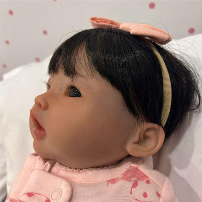 Reborn Baby Doll with white vinyl skin, brown eyes, and black hair, wearing a pink outfit with a bow headband.
