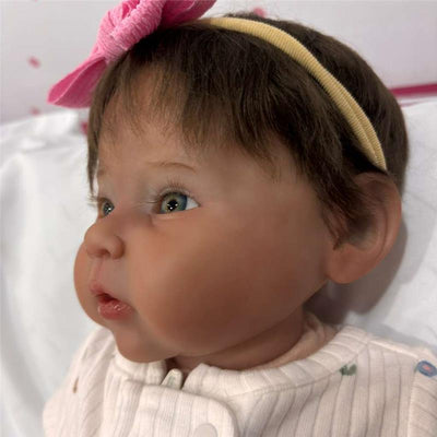 Reborn baby doll featuring white vinyl, dark brown hair, blue eyes, and a pink bow headband.