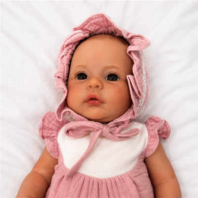 Reborn baby doll named Jocy with white vinyl, featuring a pink dress and matching bonnet, resting on a white background.
