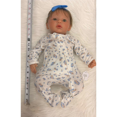 Reborn baby doll wearing a floral onesie, light brown hair with a blue bow, lying on a soft surface with a measuring tape beside.