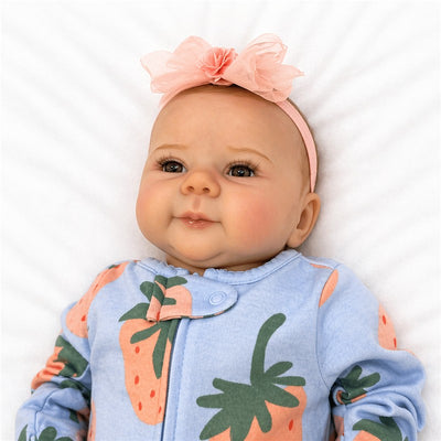 Reborn baby doll dressed in a blue outfit with strawberry prints and a pink headband, lying on a white background.