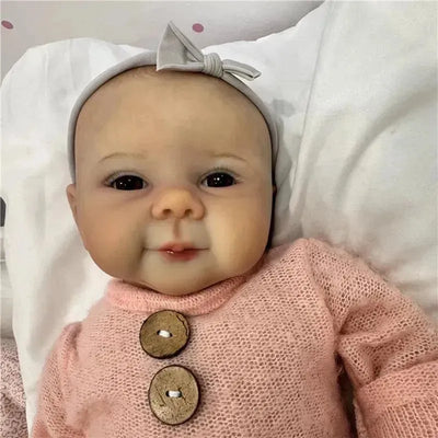 Reborn baby doll with realistic facial features, wearing a pink sweater and a gray headband, displayed on a soft background.
