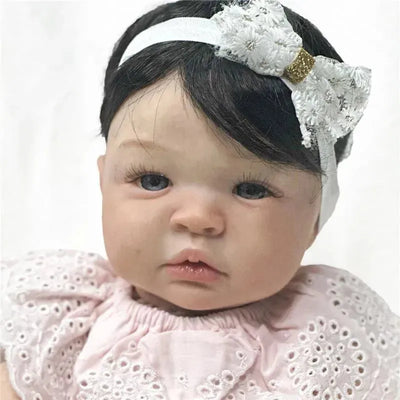 Reborn baby doll Shyann with a black wig, light pink dress, and white headband featuring a bow, showcasing realistic features.