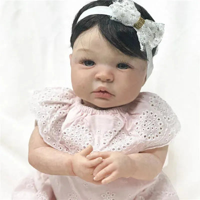 Reborn baby doll Shyann with white vinyl skin, black wig, and a pink dress featuring embroidered details.