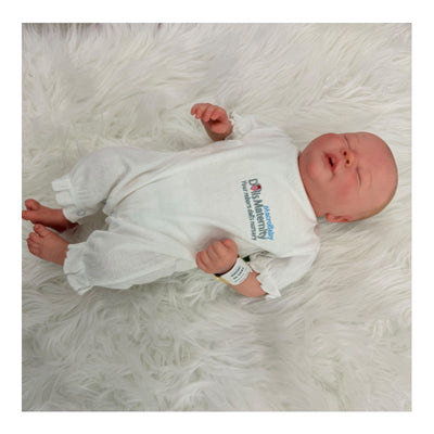 Reborn baby doll made of white vinyl, wearing a white outfit with a hospital wristband, lying on a fluffy surface.