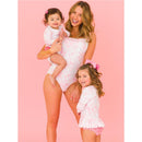 A woman holds a baby and stands beside a girl, all wearing matching pink butterfly-themed swimsuits against a pink background.