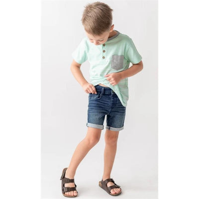 Baby boy wearing a mint green henley shirt and medium wash denim skort, adjusting his waistband while standing.