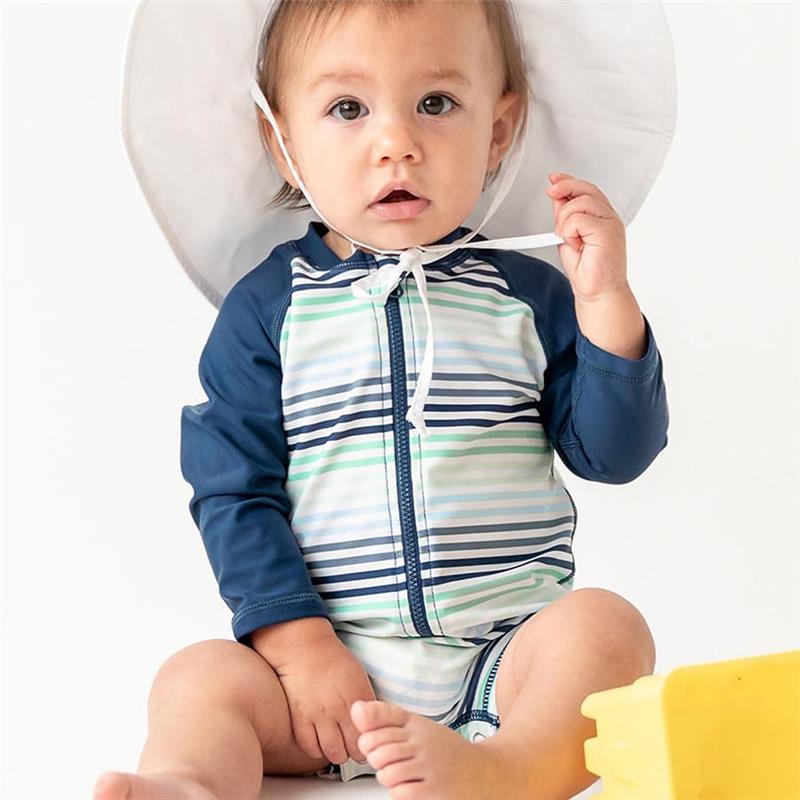 Baby boy wearing a striped long sleeve one-piece swim suit with a white sun hat, sitting on the floor.