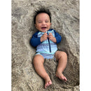 Baby boy lying in sand, wearing a striped long sleeve one-piece swimsuit with a zipper, smiling joyfully.