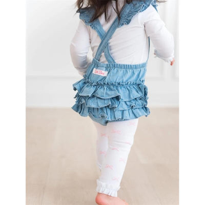 Baby girl wearing a light wash flutter overall rumper with ruffled layers, paired with white leggings featuring pink bows.