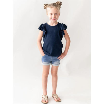 Girl smiling and posing in light wash denim shorts and a navy blue ruffle-sleeve shirt, sandals on her feet.