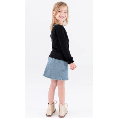 Baby girl wearing a light wash ruffle denim skirt and a black long-sleeve top, smiling while standing sideways.