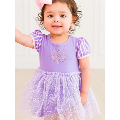 Baby girl in a purple romper with striped sleeves and a tulle skirt, smiling and raising her hand.