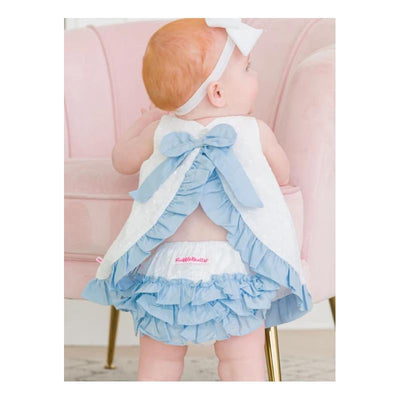 Baby girl wearing a white and light blue swing top with a bow, paired with a ruffled bloomer set, seated on a pink chair.