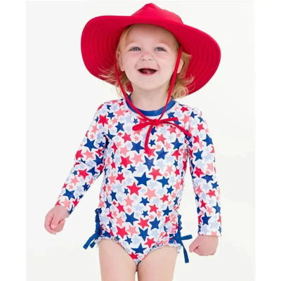 A smiling toddler in a star-patterned long sleeve rash guard with a red sun hat, ready for summer fun.