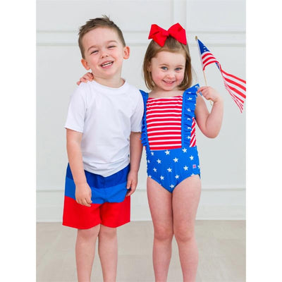 Two children, a boy in a white shirt and red shorts, and a girl in a red, white, and blue pinafore swimsuit with a bow, holding a small American flag.