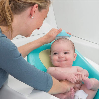 A mother bathes a smiling baby using a blue and green infant bath support in a white tub.
