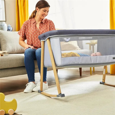 A woman sits beside a Safety 1st Rest-and-Romp 2-in-1 Bassinet & Play Yard, smiling at a baby inside it.
