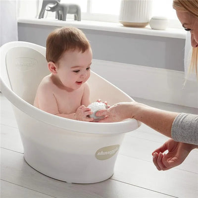 Newborn baby enjoying bath time in a white Shnuggle tub, holding a soapy bath toy while an adult helps.