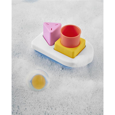 Colorful bath toy set featuring a white tray with three stacking shapes: pink triangle, orange cylinder, and yellow square, surrounded by bubbles.