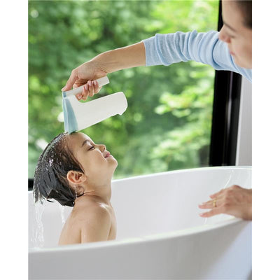 A child in a bathtub smiles as a caregiver pours water from a Skip Hop Baby Bath Rinse Cup over their head.