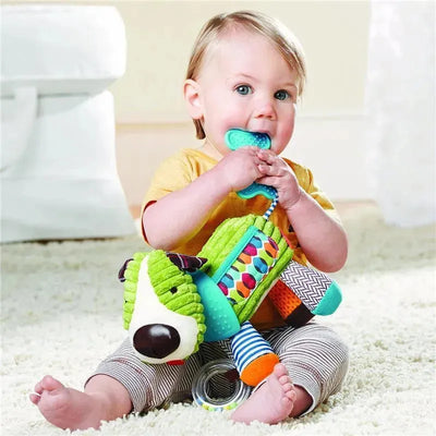 Toddler sits on a carpet holding the Skip Hop Bandana Buddies Dog stroller toy, chewing on its handle while looking at the camera.