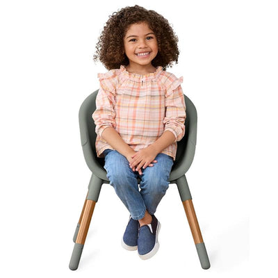 A smiling child with curly hair sits in the Skip Hop EON 4-in-1 Convertible High Chair, wearing a pastel plaid top and blue jeans.